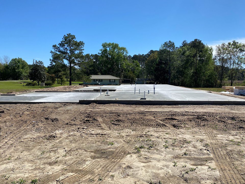 Partially built house framed with exposed wood, concrete slab foundation, dirt lot, and surrounding green trees under blue sky