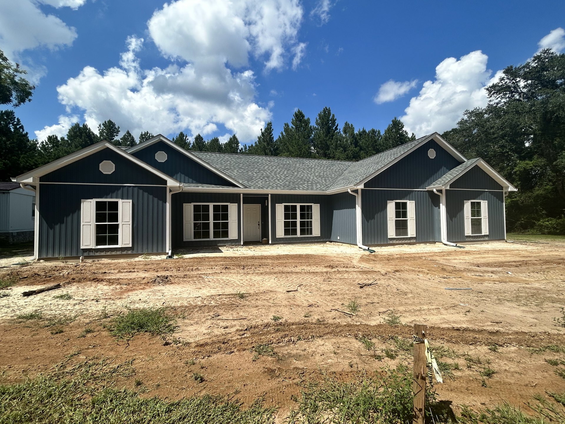 Wood-framed house under construction with white door and windows, dirt patch in front, group of trees and blue sky with clouds in background