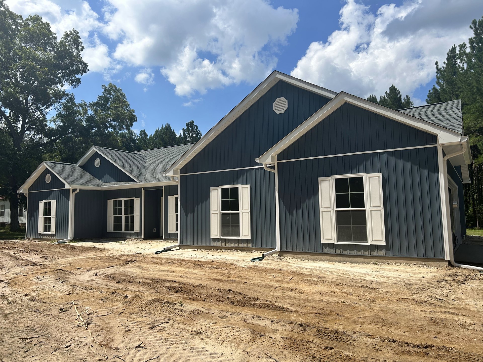 Partially built house with blue siding and roof, white-framed windows and shutters, white vent, surrounded by dirt patch, trees, and a small white cottage in the background.