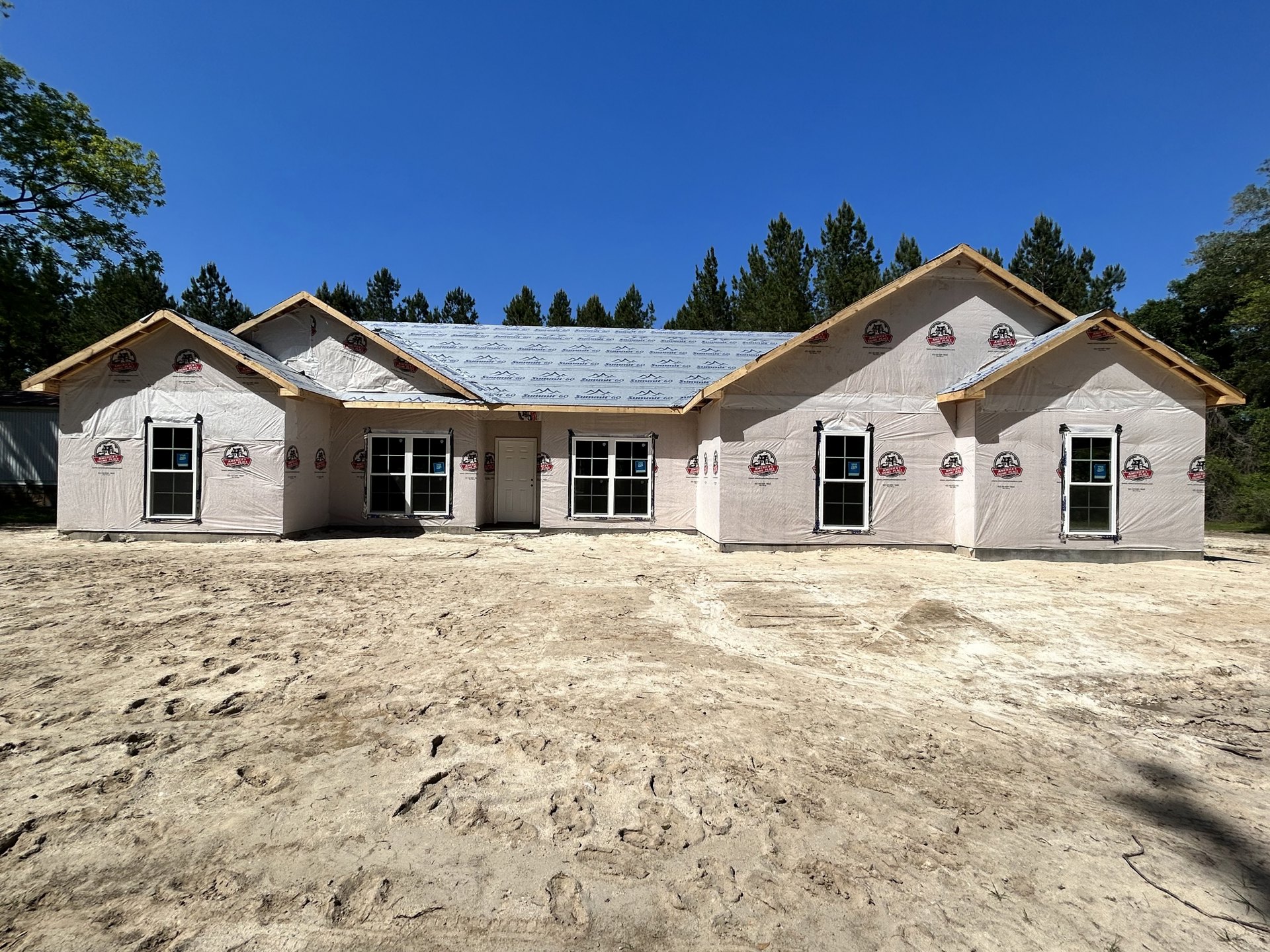 Framed house under construction with exposed plywood walls, white door and window displaying signs, sandy ground with footprints, row of leafy trees in background