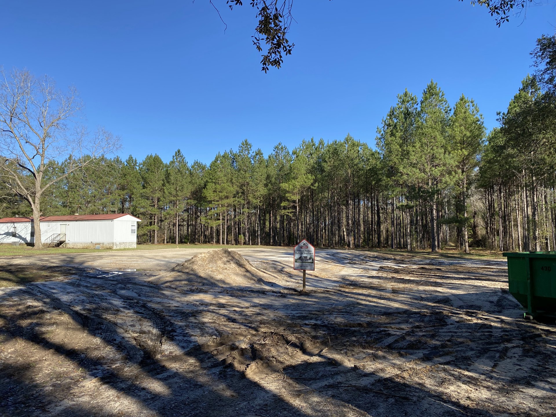 White custom home with exterior staircase, dirt road in foreground, green dumpster with numbers, construction signs, and trees under blue sky