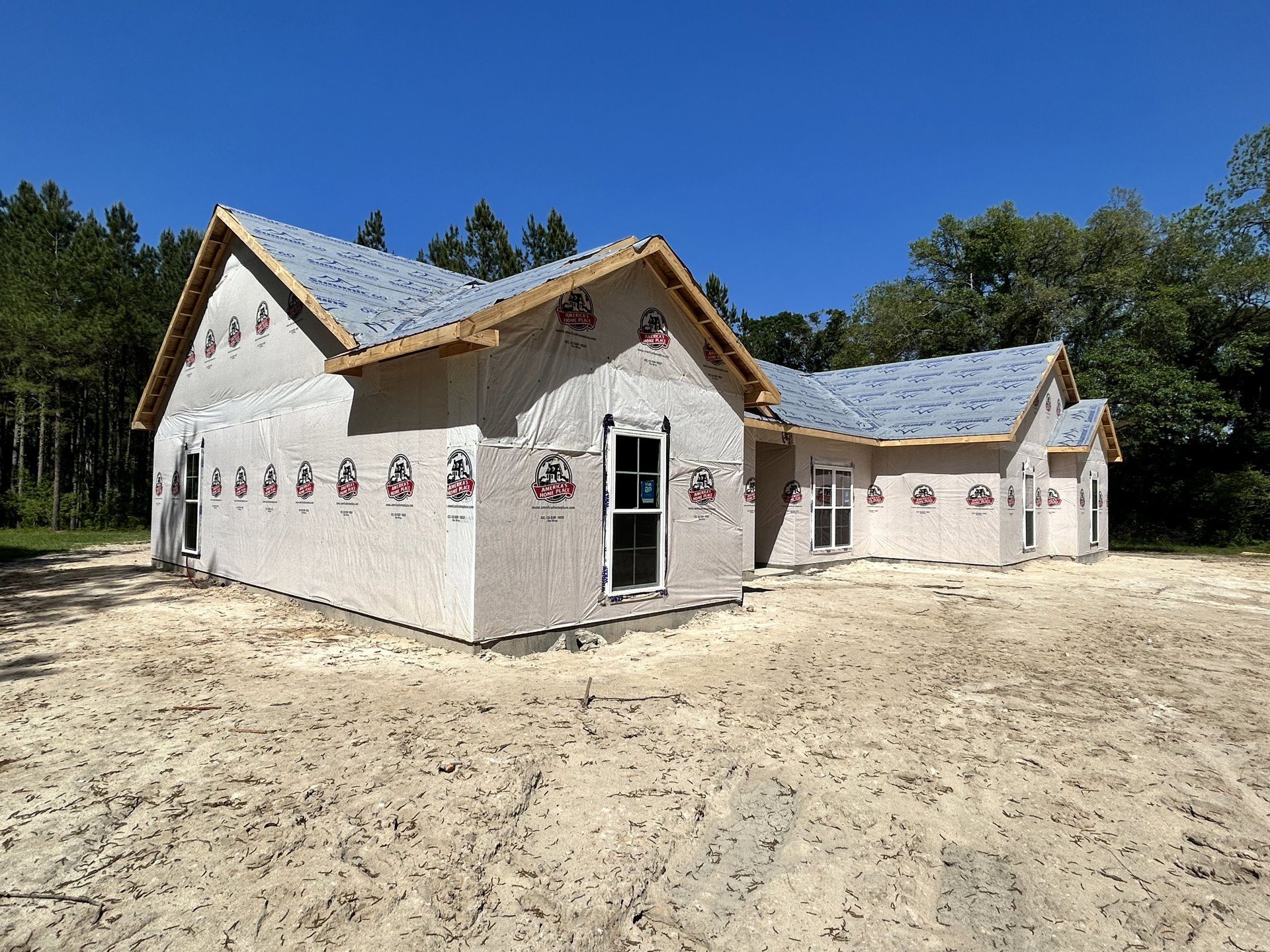 Wood-framed house under construction with plastic sheeting, sandy ground, and trees in the background; window features a sign, white surface displays a logo