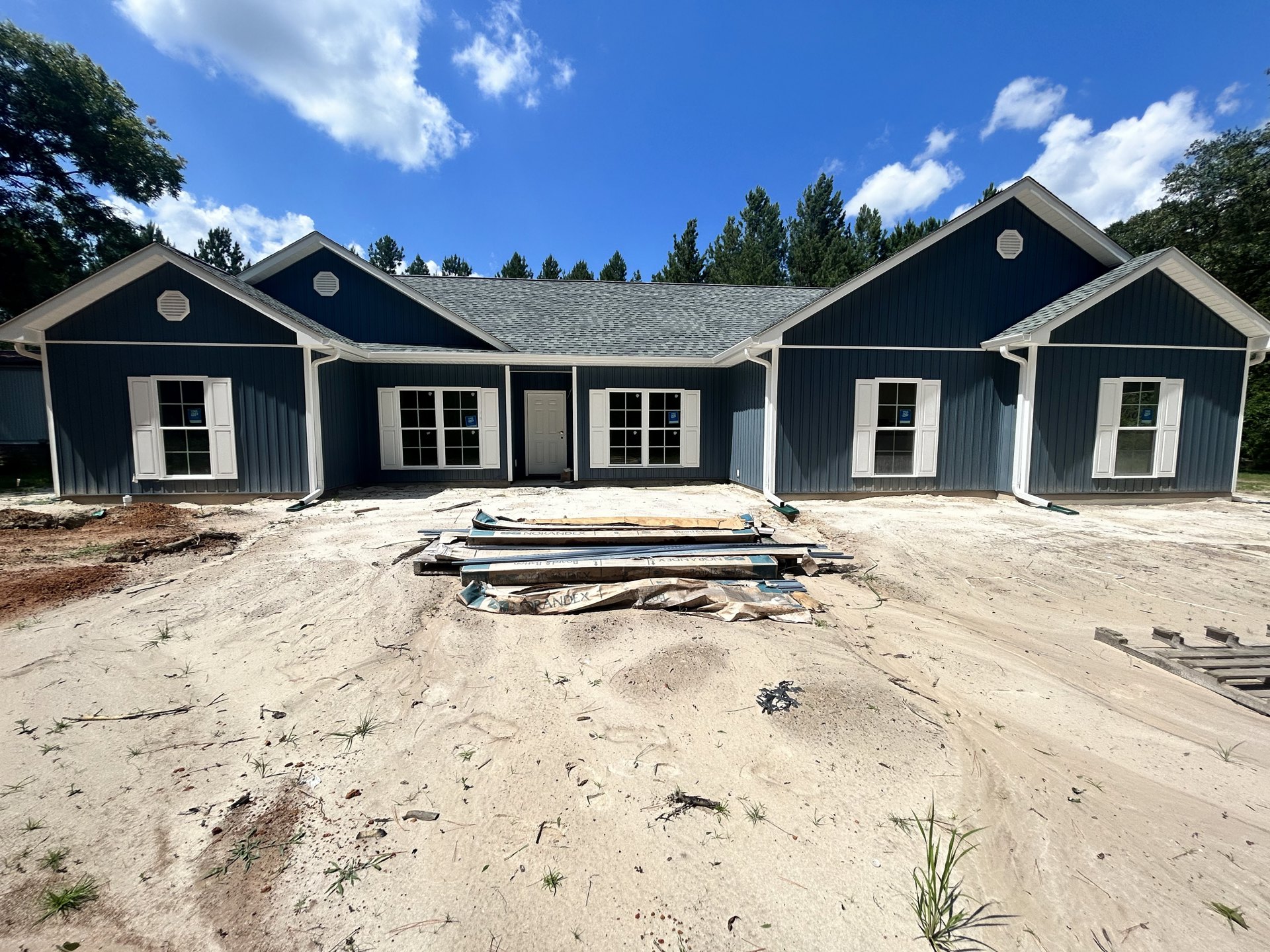Partially built house with blue roof, white trim, and white window featuring a blue sign, surrounded by dirt and mature trees under a cloudy sky