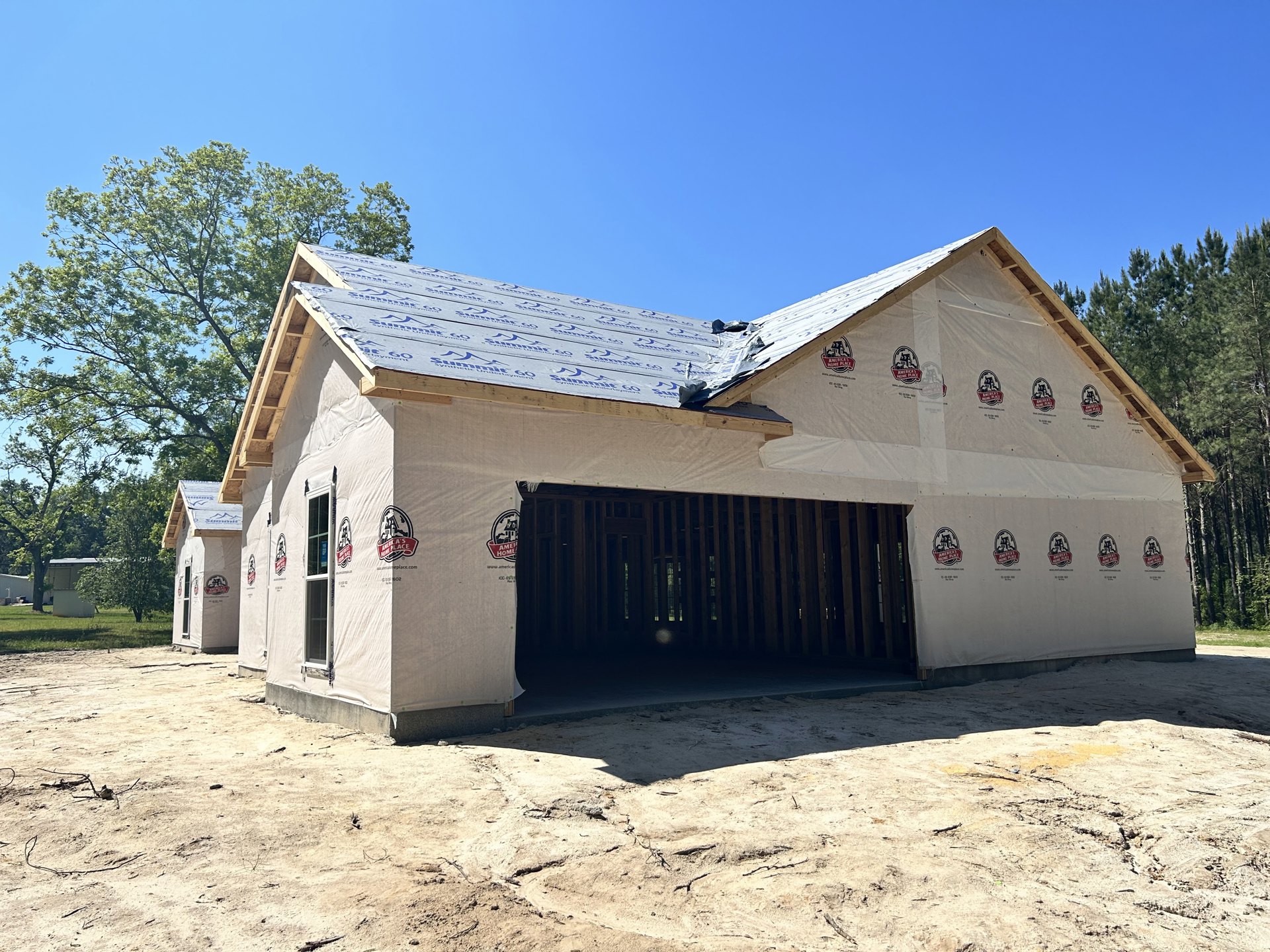 Wood-framed house under construction with attached garage, white roof panels, dirt ground covered by white sheet, large window installed, surrounded by other unfinished homes, blue
