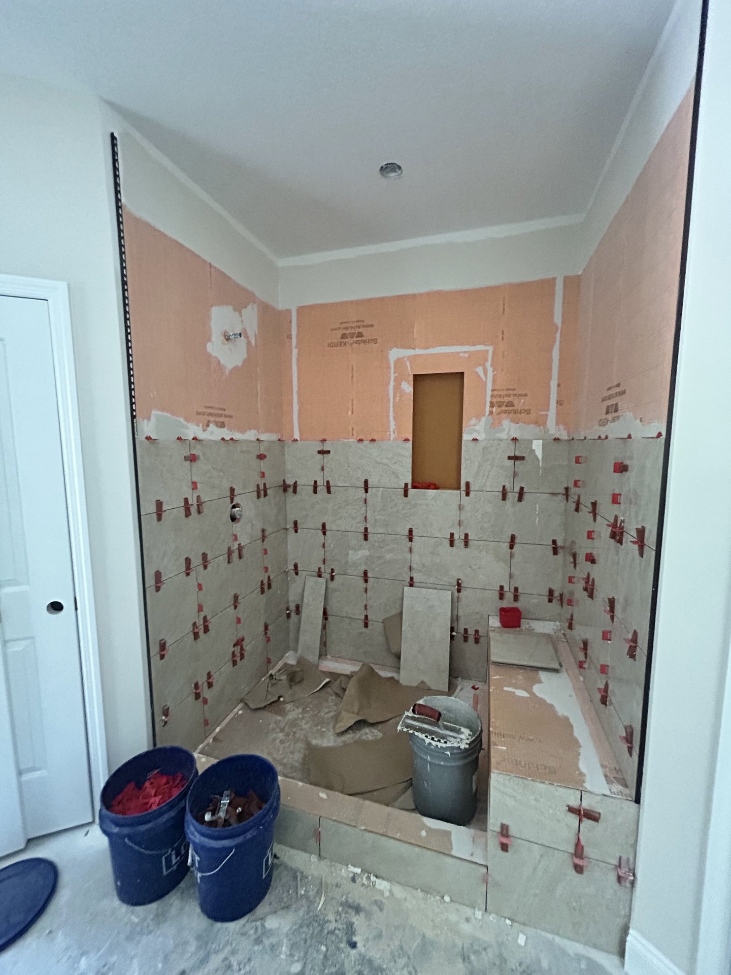 Bathroom under renovation with tiled walls, blue buckets containing tools and paint supplies, paint bucket with roller, and unfinished plaster surfaces.