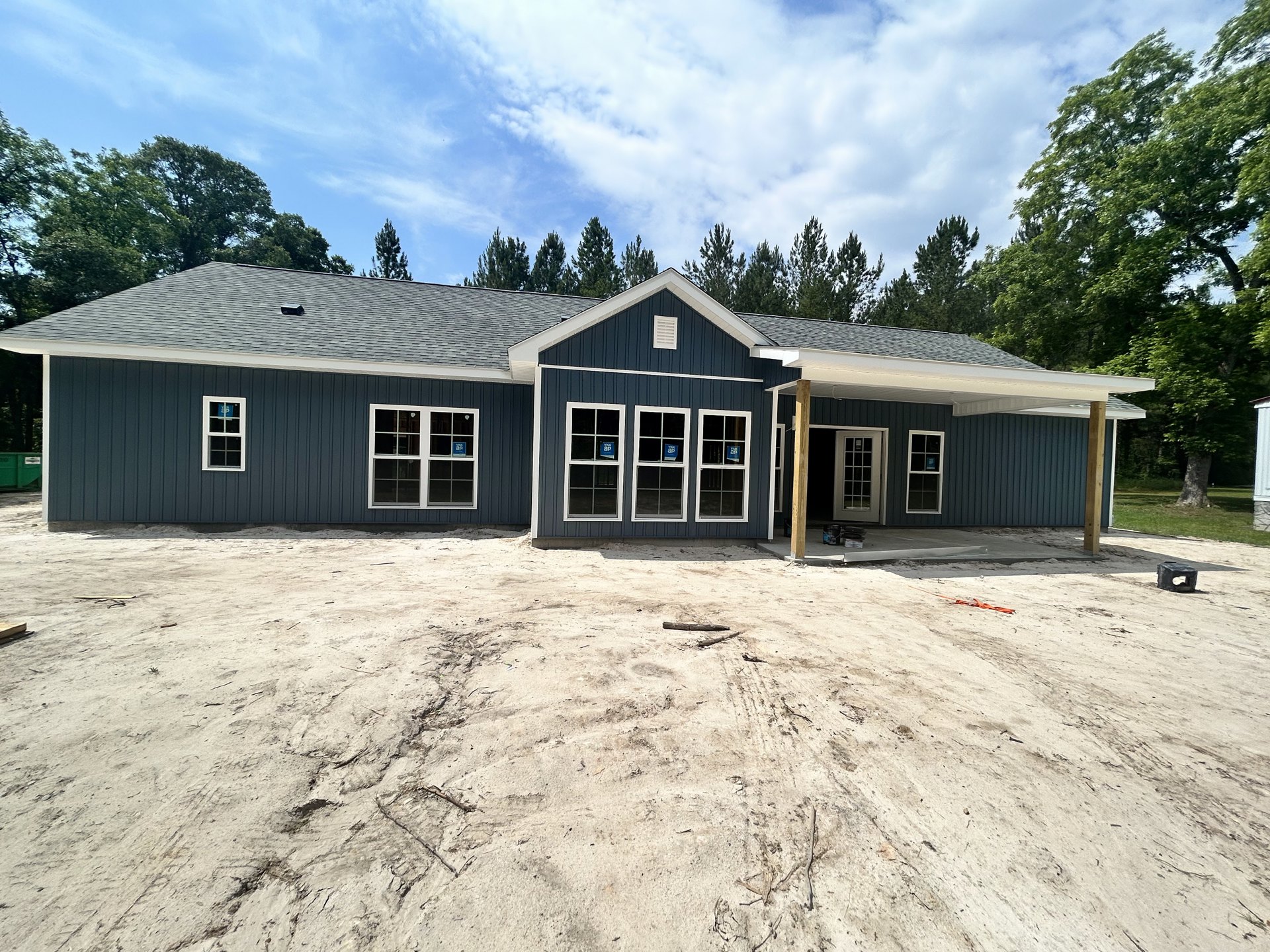 Blue house with white trim, covered porch supported by white columns, row of windows with white trim, dirt ground with tire tracks in front, window displaying a blue sign.