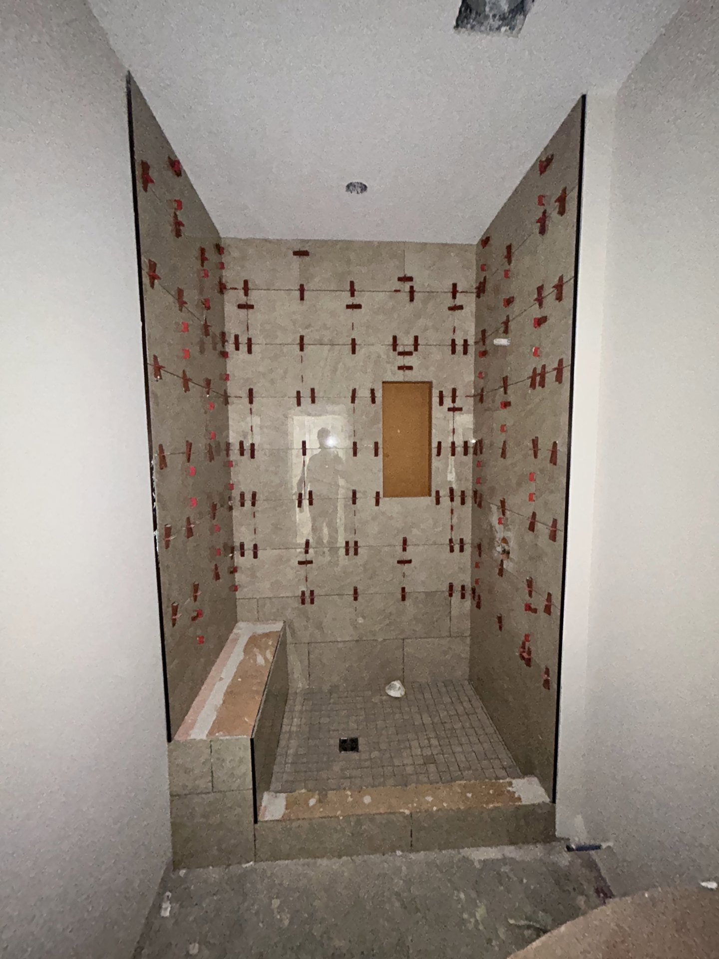 Shower with light tile walls, red tape marking sections, floor drain, and partial view of brown rectangular object with white border.