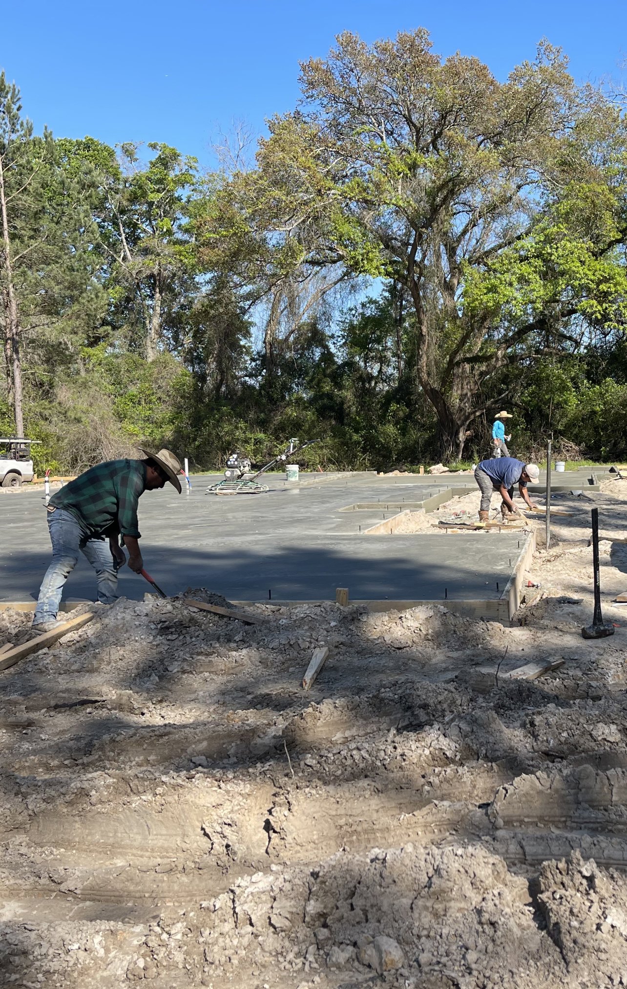 Men building a custom home on a dirt lot, one wearing a cowboy hat and hammering, another in a blue shirt and cap, white truck with black metal bars parked nearby, surrounded by