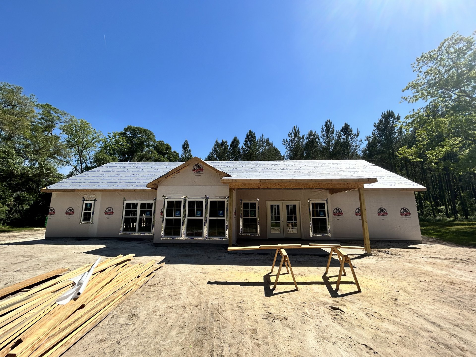 Partially built house with exposed wooden framing, roof installed, swing set in yard, sawhorse and lumber nearby, window displaying a sign, surrounded by trees under clear blue sky