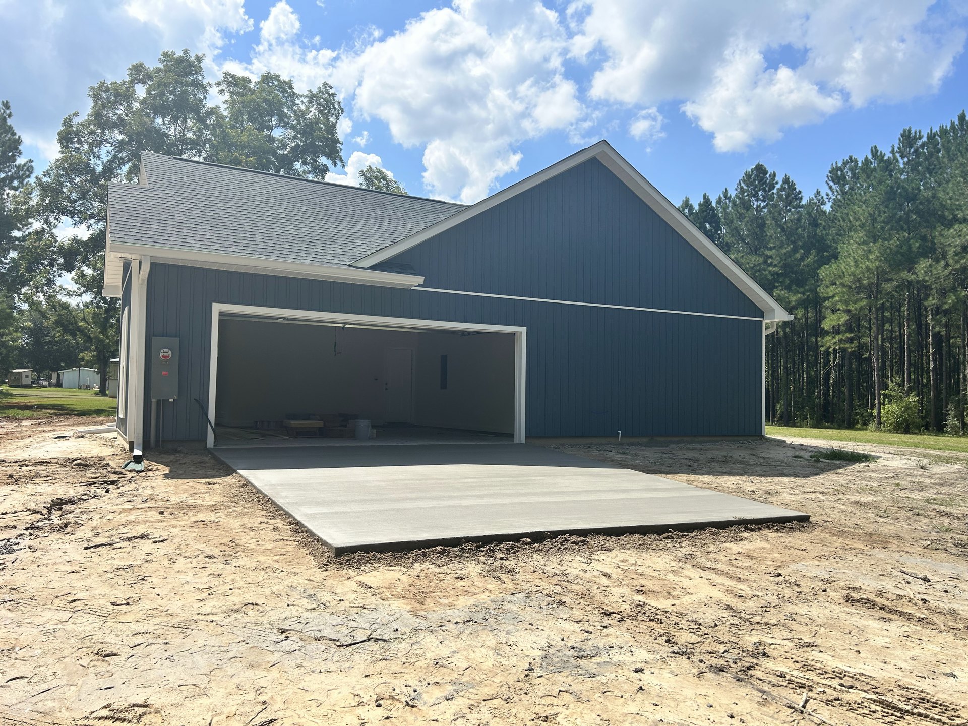 Garage with white overhead door, concrete driveway showing some dirt, trash can beside entrance, pitched roof, blue sky with scattered clouds, trees in background.