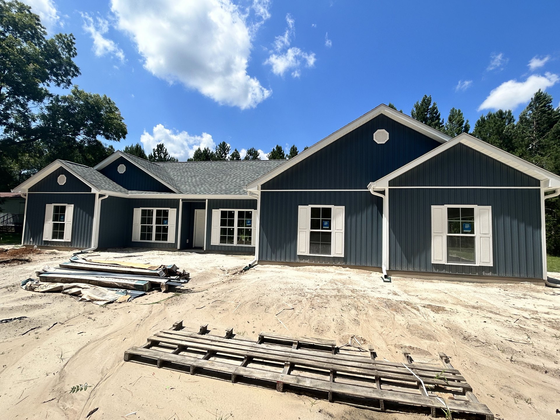 Partially built house with white-framed windows, white shutters, and a construction sign, wooden pallet resting on sandy ground, blue sky with scattered clouds and trees in