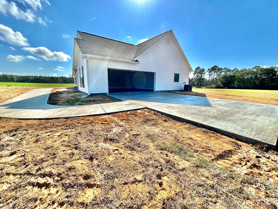 White two-car garage with paneled door, light gray siding, asphalt shingle roof, dirt driveway, and mature trees in background