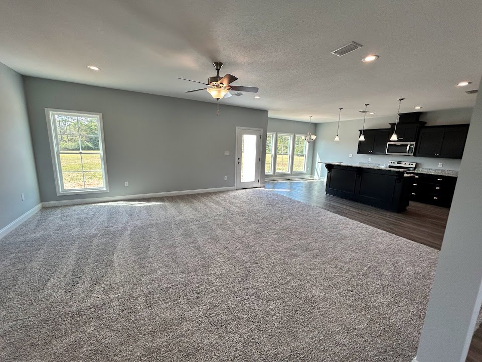 Open concept living room and kitchen with laminate flooring, black kitchen counter, white cabinetry, ceiling fan with light, carpeted area, large window overlooking grassy field