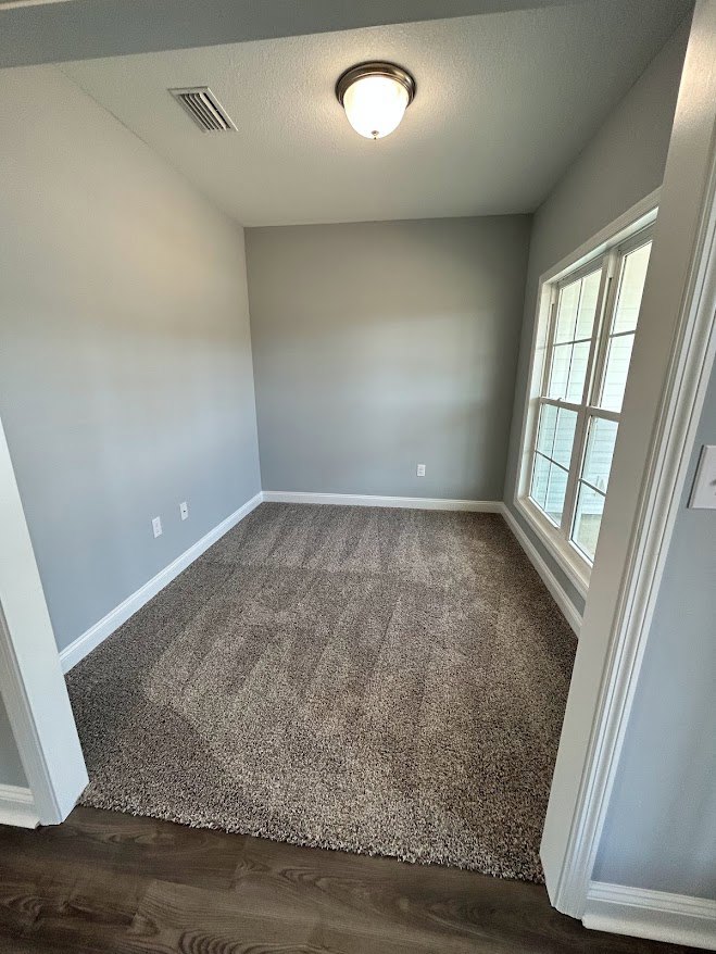 Neutral-toned carpeted room with white-framed window, ceiling light fixture, and ceiling vent, surrounded by light-colored walls and simple molding.
