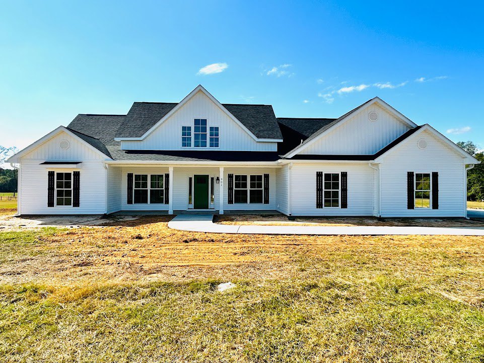 White house with green front door, black shuttered windows, white trim, concrete foundation, grassy lawn, Southfork Ranch visible in background, blue sky with scattered clouds