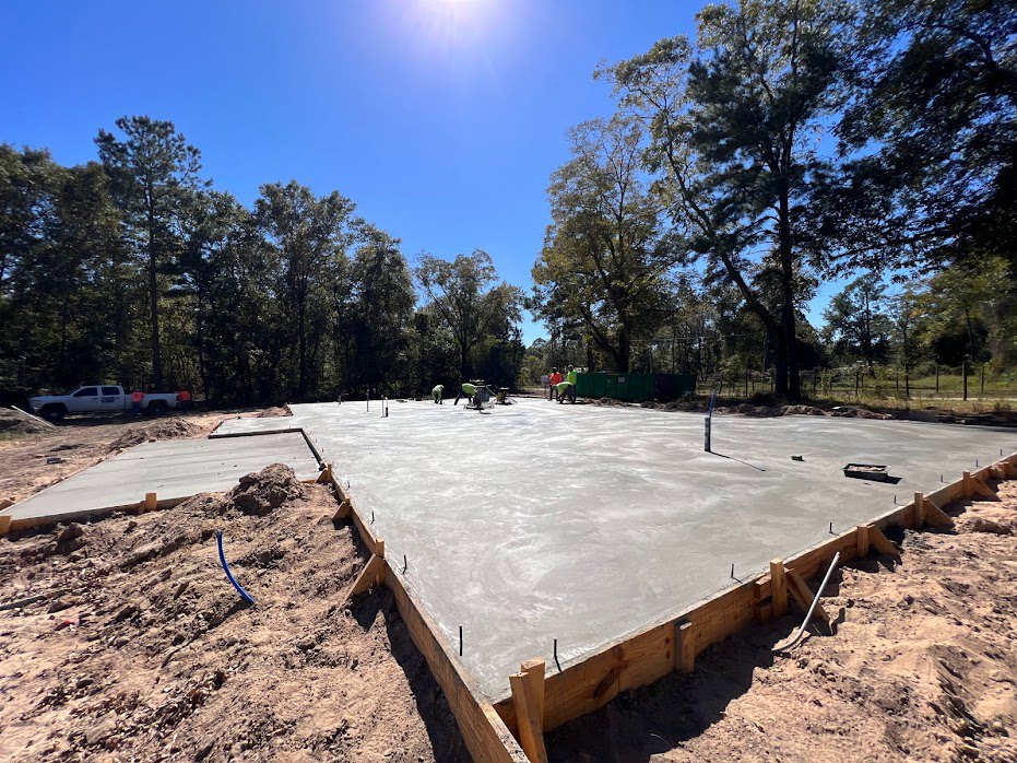 Concrete slab under construction on dirt lot, surrounded by wooden framing, with leafy trees and blue sky in the background