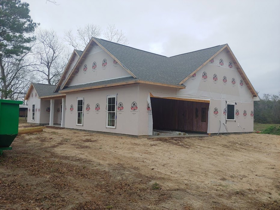 Framed house under construction with attached garage, dark wood paneling, dirt field in foreground, large tree with bare branches, green waste container, Robert Frost Farm cottage