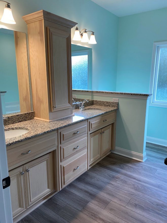 Bathroom featuring a marble countertop, rectangular mirror, three-light fixture, wooden floor with built-in drawers, blue and white window, white walls with wooden trim