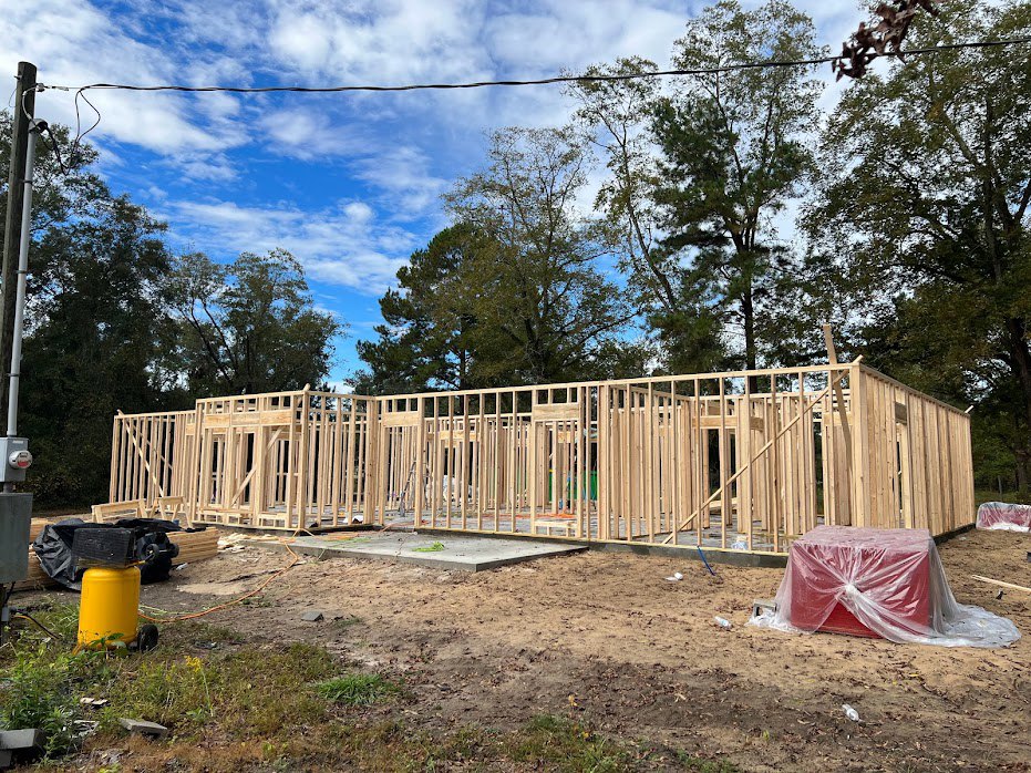 Wood-framed house under construction with exposed beams, red plastic-wrapped box, yellow cylinder with black mesh, surrounding trees, and blue sky