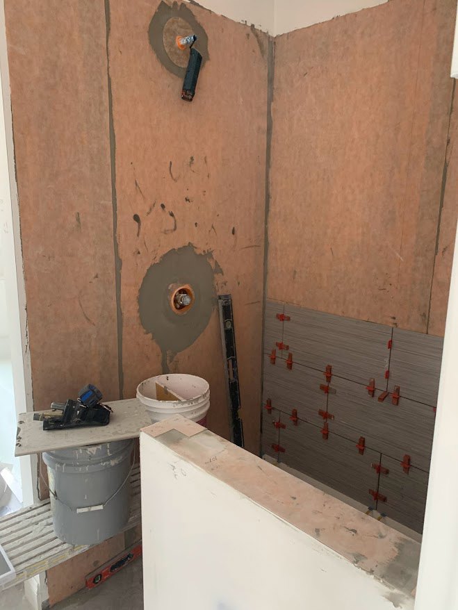 Unfinished bathroom with white plaster walls, exposed plumbing, red wall pegs, white bucket containing wood, and construction tools on tiled floor