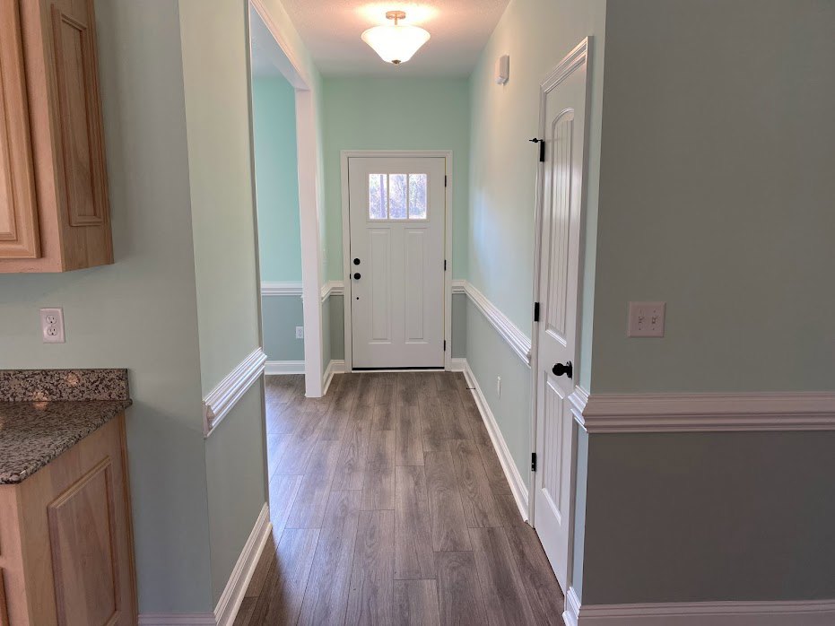 Hallway with light wood flooring, white door with glass window, white walls, ceiling light fixture, partial view of kitchen countertop and cabinetry