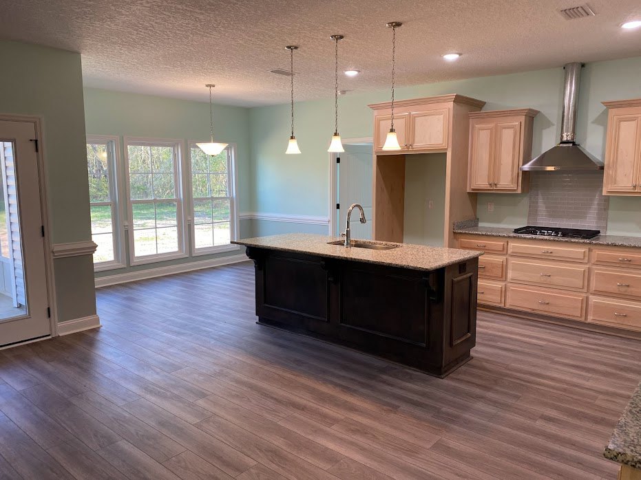 Modern kitchen featuring a central island with white countertop, wood cabinetry, stainless steel sink, green accent wall, silver range hood, wood flooring, large windows, and a