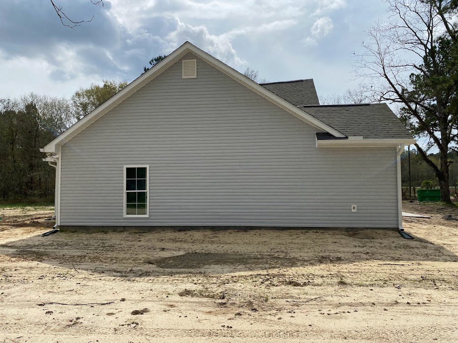 White-sided house with attached garage, white-framed window, metal wall bordering dirt field, tree near exterior, white roof with trees in background.