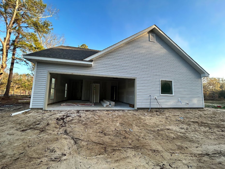 Partially built house with light siding, attached garage, circular window, stack of lumber, dirt ground with tire tracks, and mature trees in the background