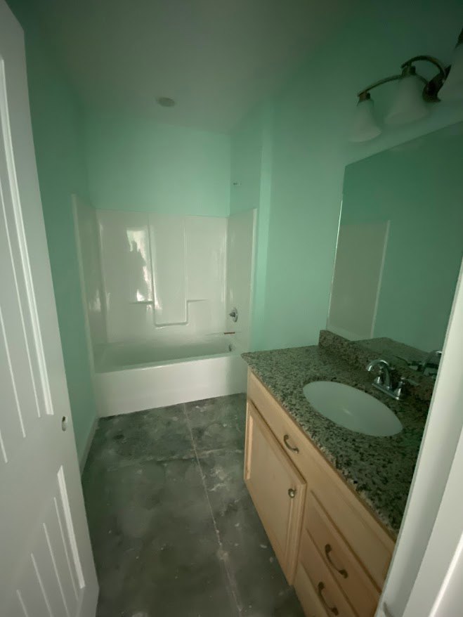 White oval sink set in a speckled granite countertop, freestanding bathtub with chrome fixtures, light gray tile flooring, wood cabinetry, large wall mirror, and neutral painted