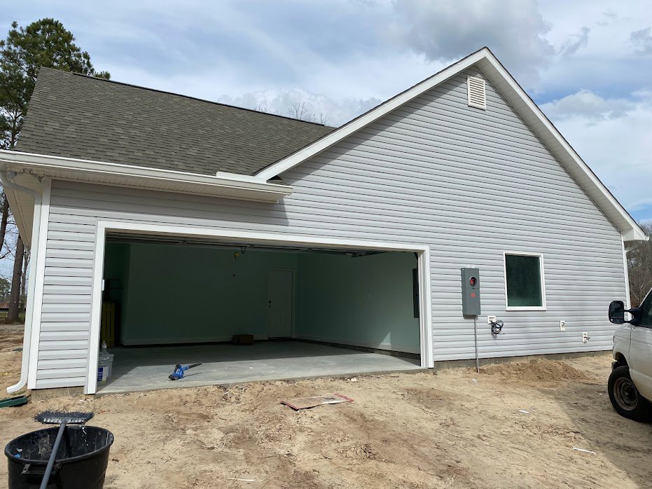 Garage with large open door, white truck parked inside, white-framed window on siding, black bucket and long pole near entrance, cloudy sky above.