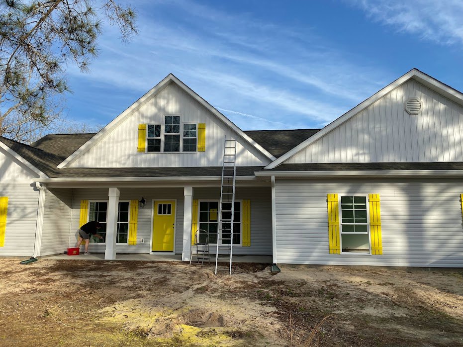 Man in shorts and black shirt painting house exterior near yellow door with black trim and window with yellow shutters; ladder leaning against siding on porch.
