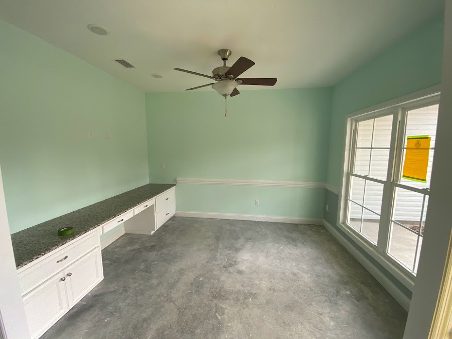 Ceiling fan with light fixture, large windows with yellow sign taped to glass, grey flooring, white plaster walls, roll of tape on countertop, built-in cabinetry
