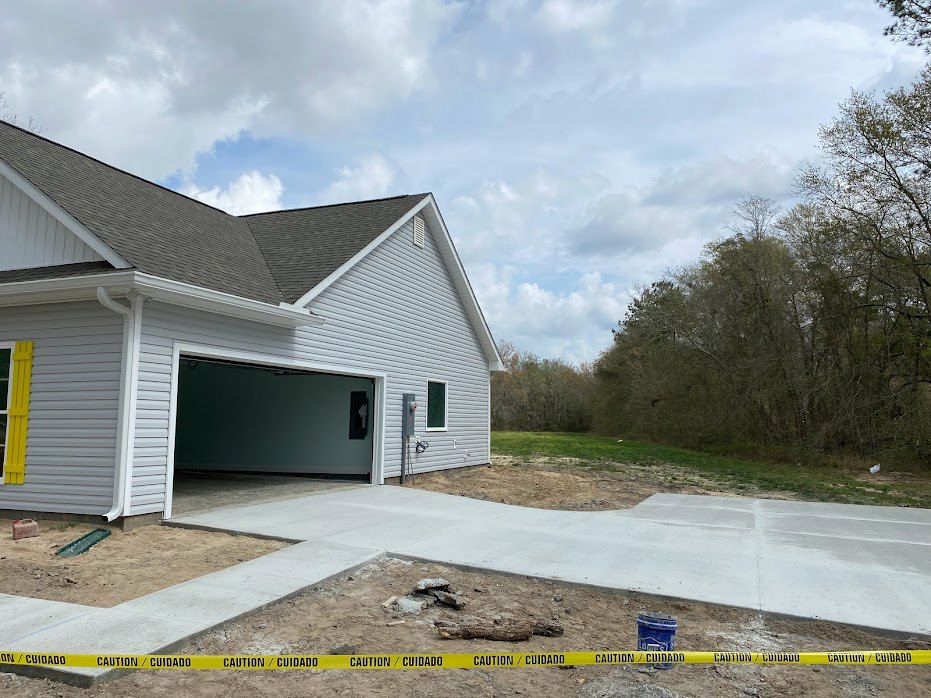 Two-story home with light-colored siding, attached garage with white door, concrete driveway bordered by rocks, grassy lawn, mature trees, and partly cloudy blue sky