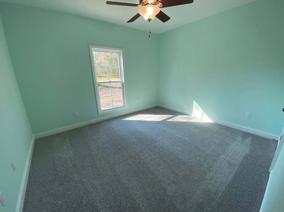 Carpeted room with white walls, ceiling fan with light fixture, large window facing fenced backyard