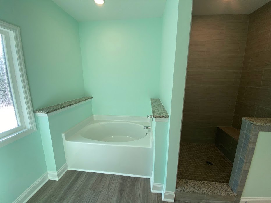 White freestanding bathtub beside glass-enclosed shower, wood plank flooring, white tile walls, chrome fixtures, and a white door frame.