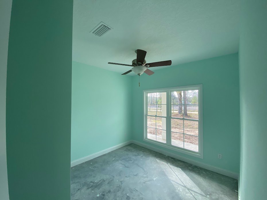 Ceiling fan with light fixture mounted on white plaster ceiling, large window showing leafy trees outside, light blue flooring, wall vent near fan