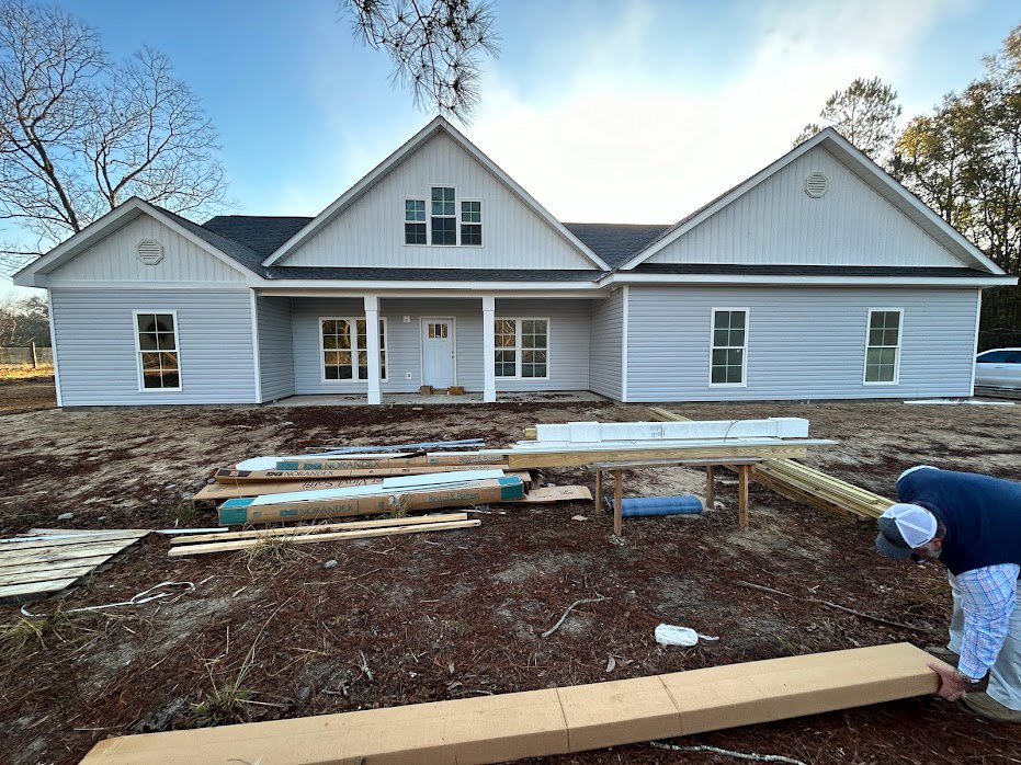 Modern home exterior with wood siding, large windows with white frames, front porch, and stacked lumber in the yard.