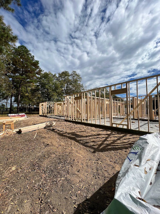 Wood-framed house under construction on dirt lot, surrounded by trees and blue sky, white construction bag on ground, wooden planks and temporary fencing visible