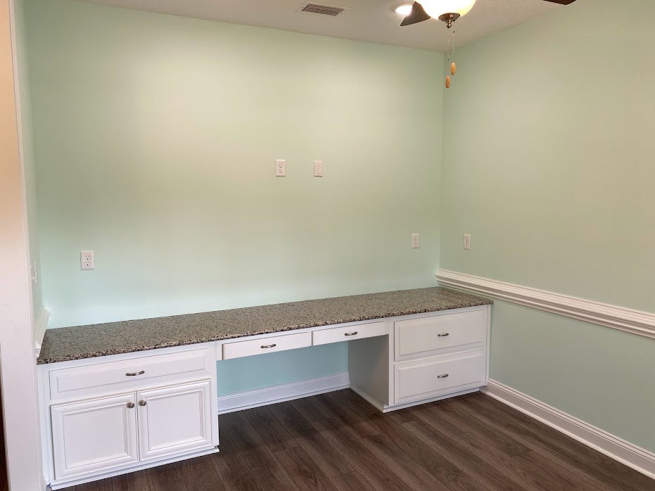 Wood floor with white trim, white kitchen cabinetry and drawers, countertop, ceiling fan with light fixture, desk against wall with white molding