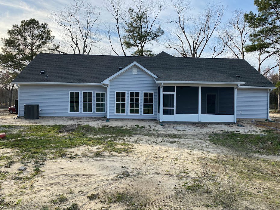 Modern house with light-colored siding, several rectangular windows, and a dirt yard with visible tire tracks in the foreground