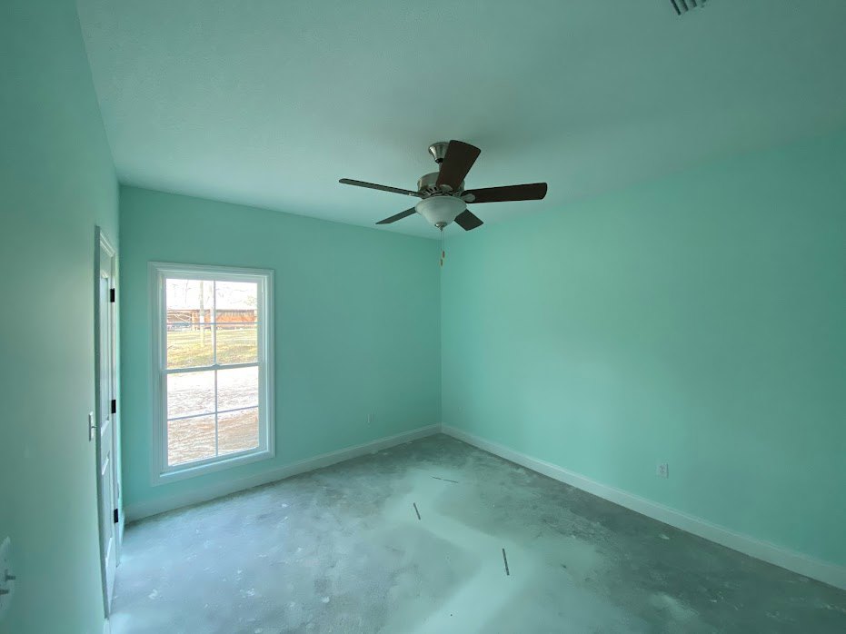 Concrete floor and white plaster walls in a room with a ceiling fan and light fixture, large window overlooking a grassy field.