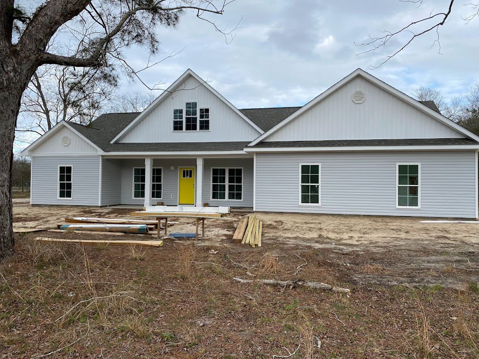 White cottage-style home with yellow front door, multi-pane windows, gray siding, and a pile of stacked wood near the entrance, surrounded by trees under a clear sky