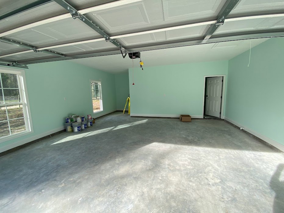 Garage interior with white overhead door, paint cans grouped on concrete floor, white ceiling with garage door opener, window showing fenced yard, plaster walls.