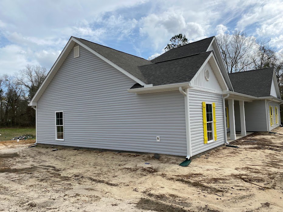 Partially built house with exposed framing, yellow-trimmed window, gray roof, and dirt lot surrounded by trees under cloudy sky