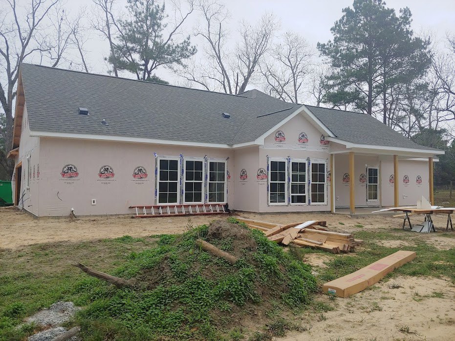 Two-story house under construction with exposed framing, shingled roof, installed windows with white frames, pile of lumber in front yard, brown curb edging grassy area, and dirt