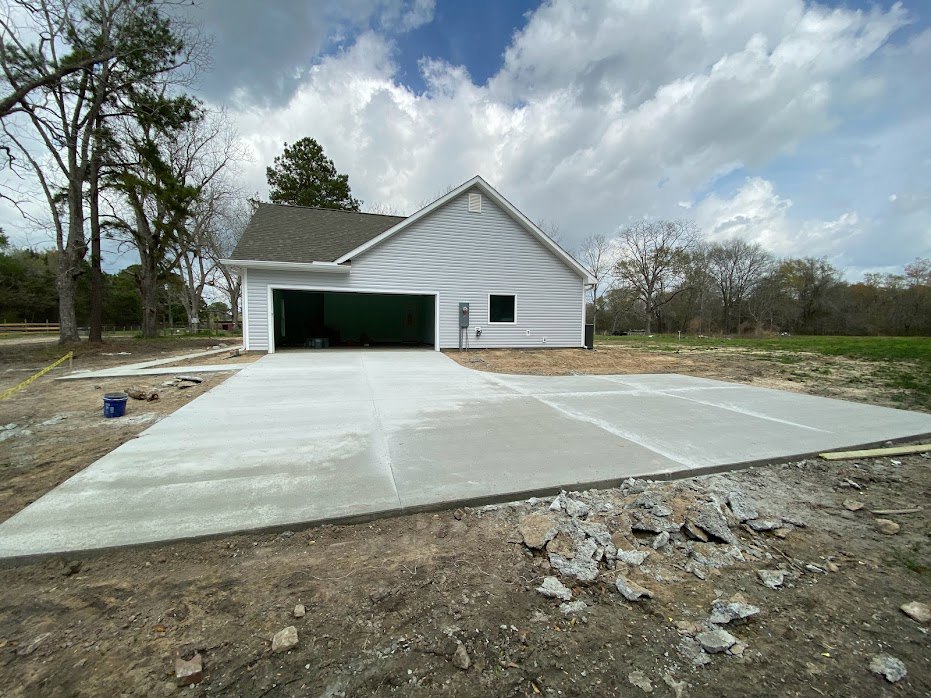 Two-story house with attached garage, concrete driveway, light siding, and gabled roof under blue sky with scattered clouds, surrounded by mature trees