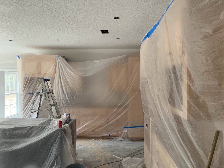 Room under renovation with plastic sheets covering walls, aluminum ladder with blue top, paint can on table, person partially visible behind plastic, white-framed window, plaster