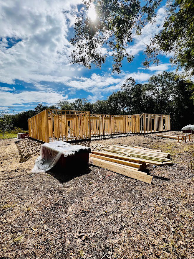 Wood-framed house under construction with exposed beams, stacks of lumber, rolls of plastic sheeting, surrounded by trees beneath a clear blue sky