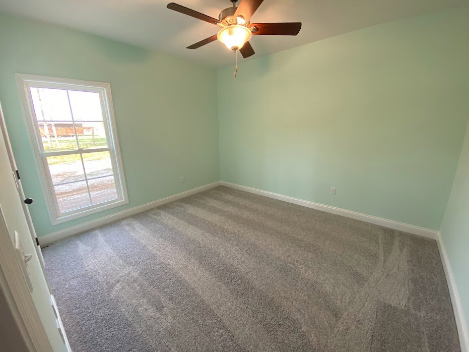 Carpeted bedroom with white walls, ceiling fan with light fixture, large window overlooking farmland.