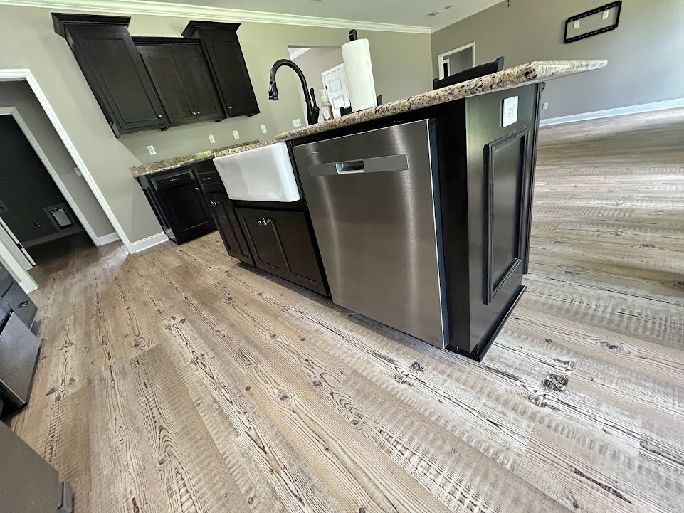 Kitchen with stainless steel sink, black skull-shaped faucet, marble-topped white island, stainless steel dishwasher, dark cabinetry, wooden floor, and framed artwork on wall